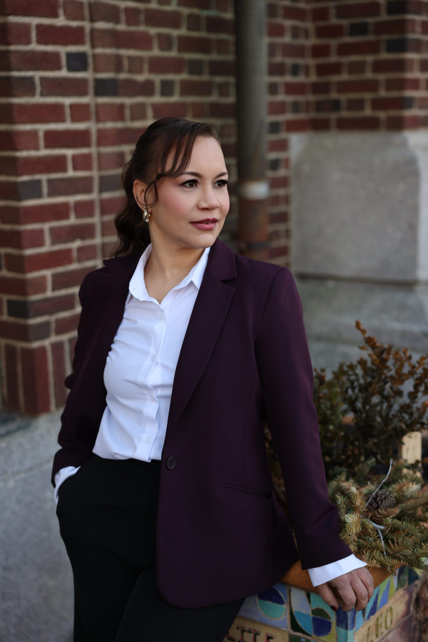 Confident woman in a burgundy blazer stands outdoors near a brick wall.