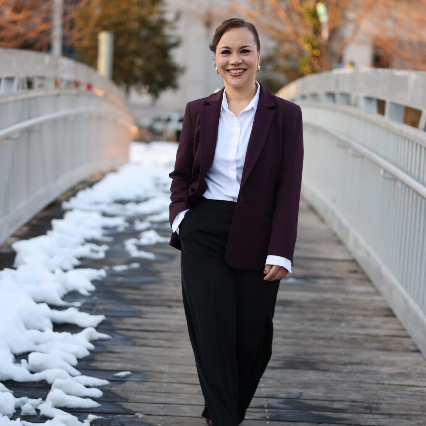 A woman in business attire stands on a snowy wooden bridge outdoors.